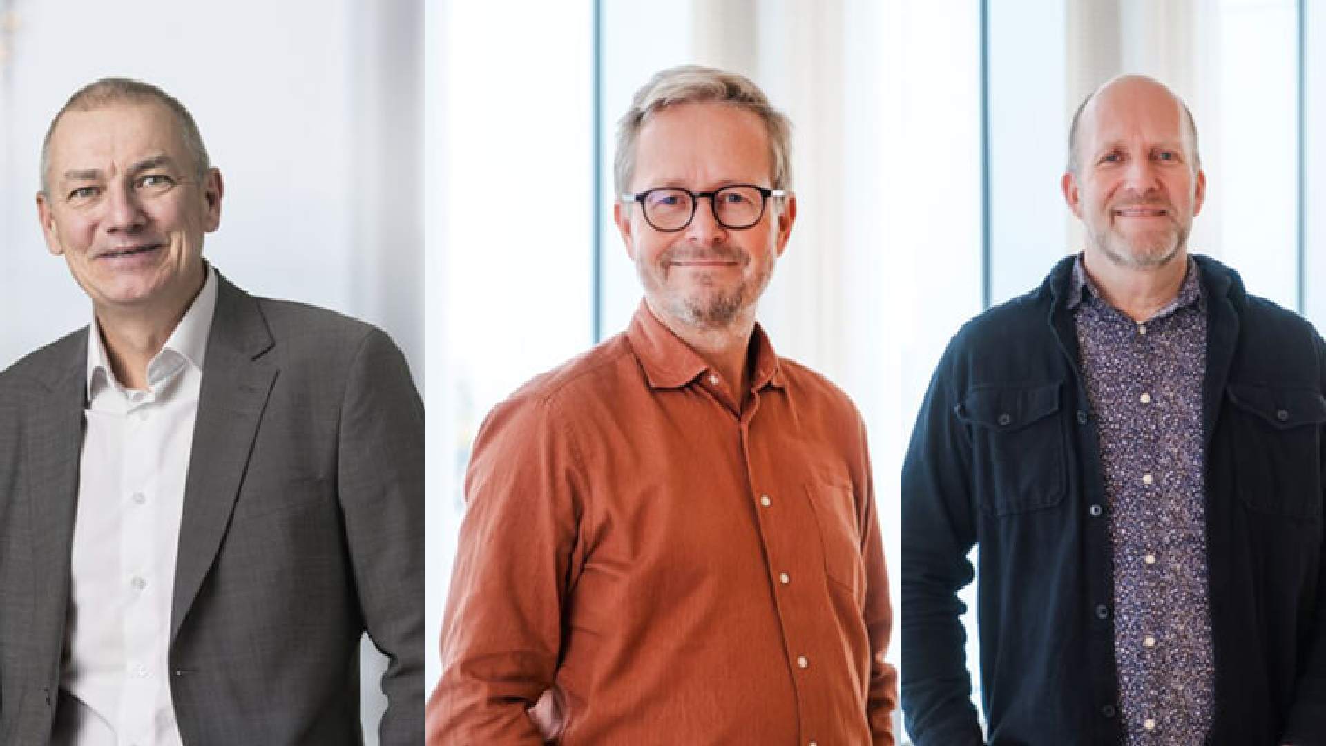 Three individuals standing indoors in front of large windows, dressed in business casual attire including a grey suit, an orange button-up shirt, and a dark jacket over a patterned shirt