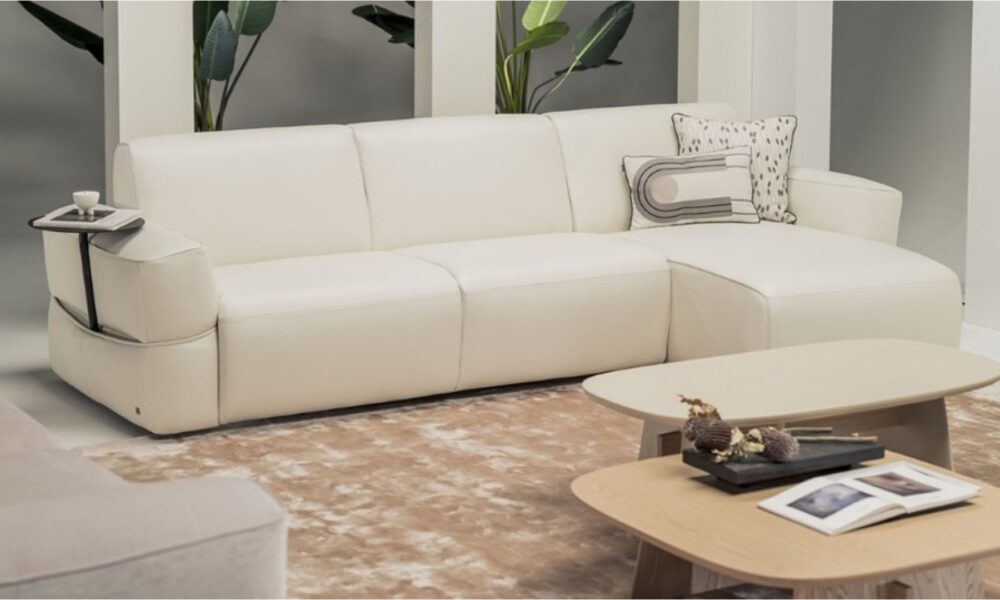 Modern white leather sectional sofa with integrated side tray, decorative cushions, and a beige rug, paired with two light wood coffee tables featuring books and dried floral accents, set in a contemporary living room with green potted plants in the background."