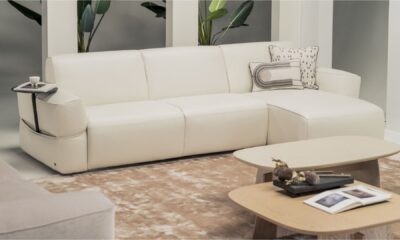 Modern white leather sectional sofa with integrated side tray, decorative cushions, and a beige rug, paired with two light wood coffee tables featuring books and dried floral accents, set in a contemporary living room with green potted plants in the background."