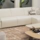 Modern white leather sectional sofa with integrated side tray, decorative cushions, and a beige rug, paired with two light wood coffee tables featuring books and dried floral accents, set in a contemporary living room with green potted plants in the background."