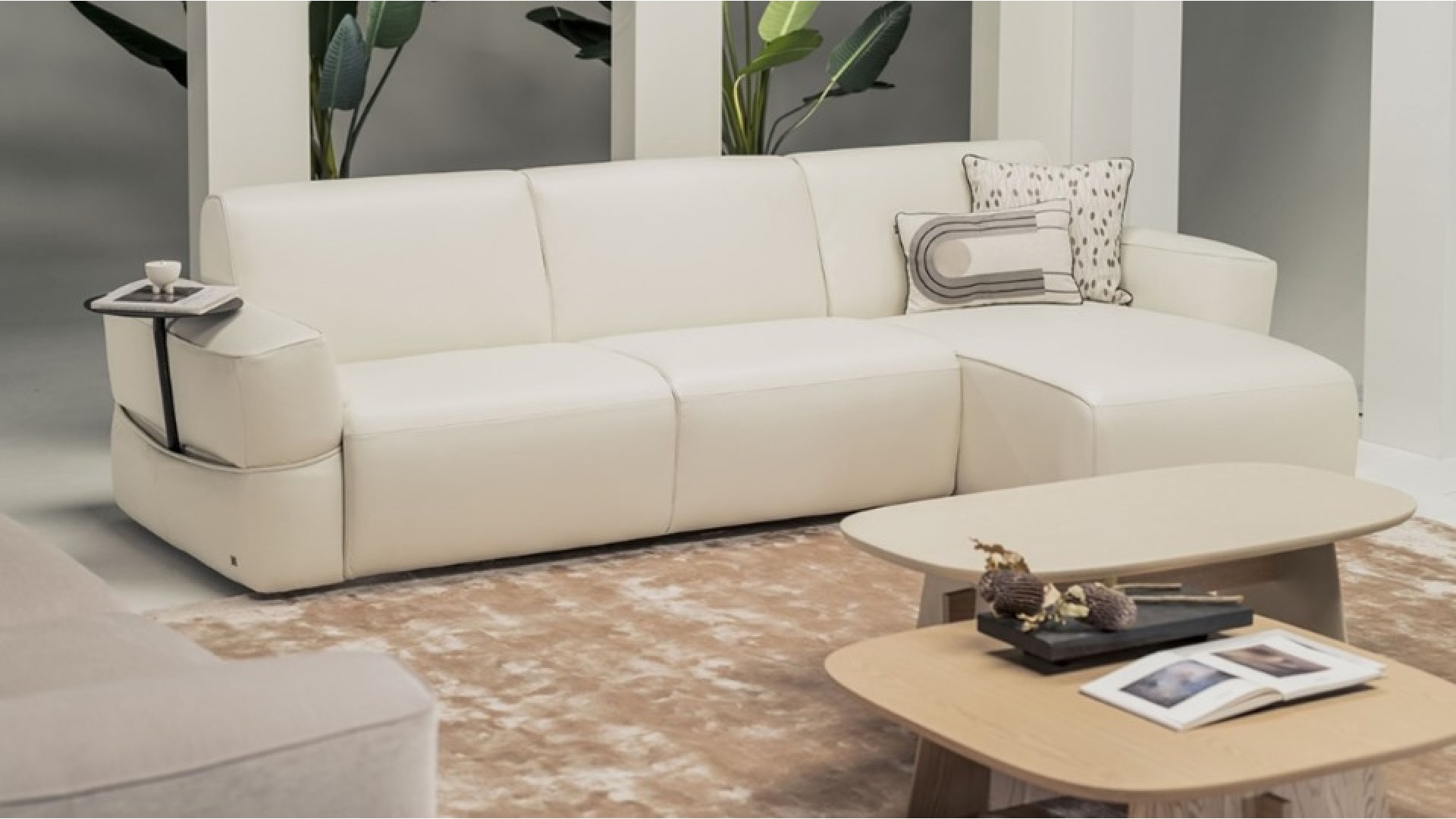Modern white leather sectional sofa with integrated side tray, decorative cushions, and a beige rug, paired with two light wood coffee tables featuring books and dried floral accents, set in a contemporary living room with green potted plants in the background."