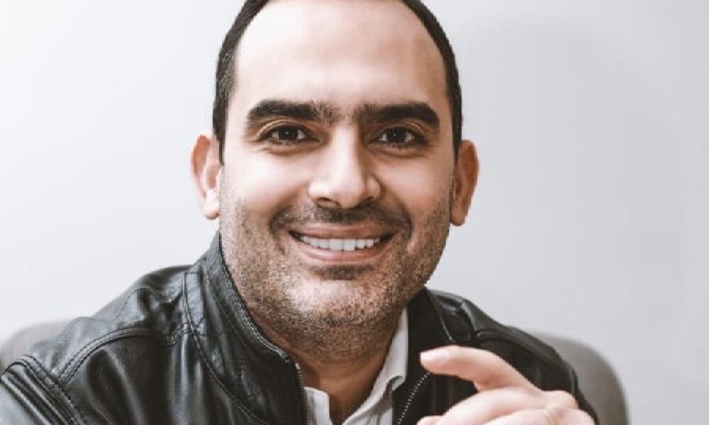 Person seated indoors wearing a black leather jacket over a white collared shirt, hands clasped together, sitting on a gray sofa against a plain light-colored wall.