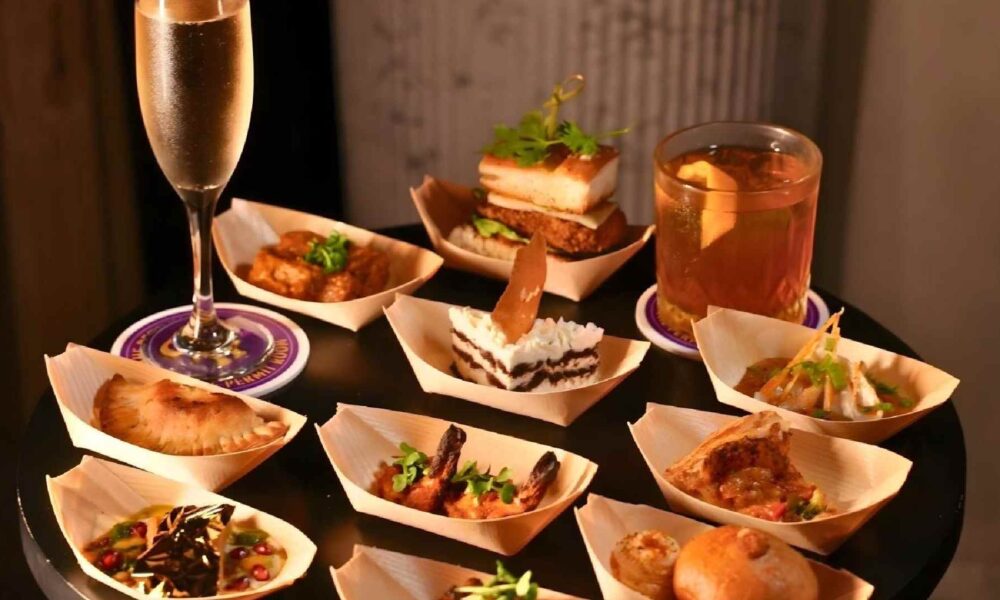 Assorted tapas-style dishes served in small wooden bowls on a round black table, featuring items like grilled prawns, stuffed pastries, bite-sized desserts, and savory snacks, accompanied by a glass of champagne and a cocktail.
