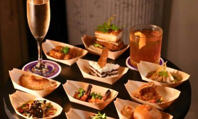 Assorted tapas-style dishes served in small wooden bowls on a round black table, featuring items like grilled prawns, stuffed pastries, bite-sized desserts, and savory snacks, accompanied by a glass of champagne and a cocktail.