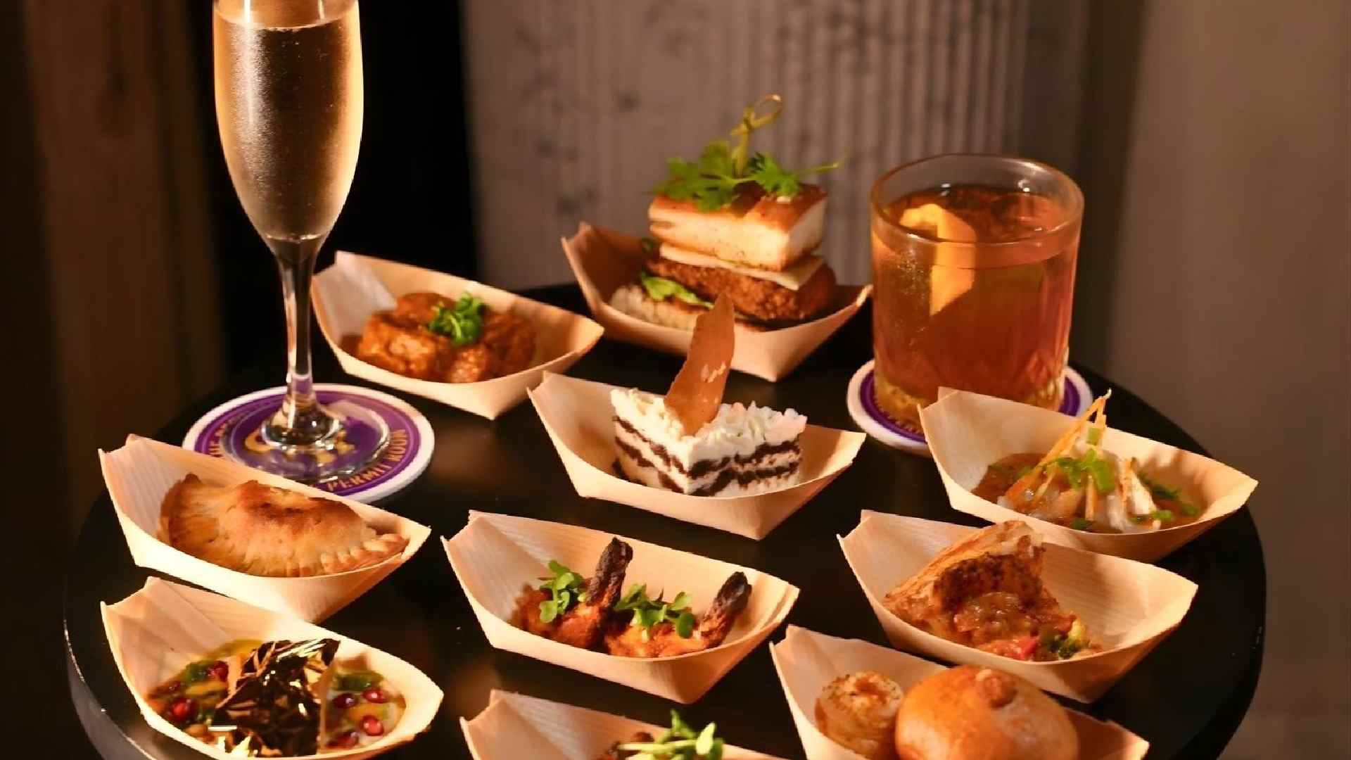 Assorted tapas-style dishes served in small wooden bowls on a round black table, featuring items like grilled prawns, stuffed pastries, bite-sized desserts, and savory snacks, accompanied by a glass of champagne and a cocktail.