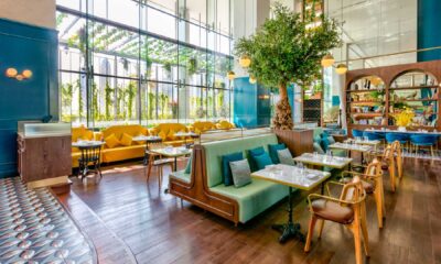 Interior of Couqley French Brasserie at Pullman Dubai Downtown featuring elegant high ceilings, large floor-to-ceiling windows with natural light, green and yellow seating booths, marble-top tables, wooden flooring, and a central decorative tree creating a vibrant upscale dining atmosphere