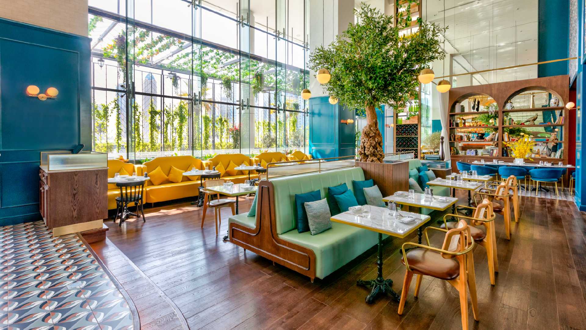 Interior of Couqley French Brasserie at Pullman Dubai Downtown featuring elegant high ceilings, large floor-to-ceiling windows with natural light, green and yellow seating booths, marble-top tables, wooden flooring, and a central decorative tree creating a vibrant upscale dining atmosphere