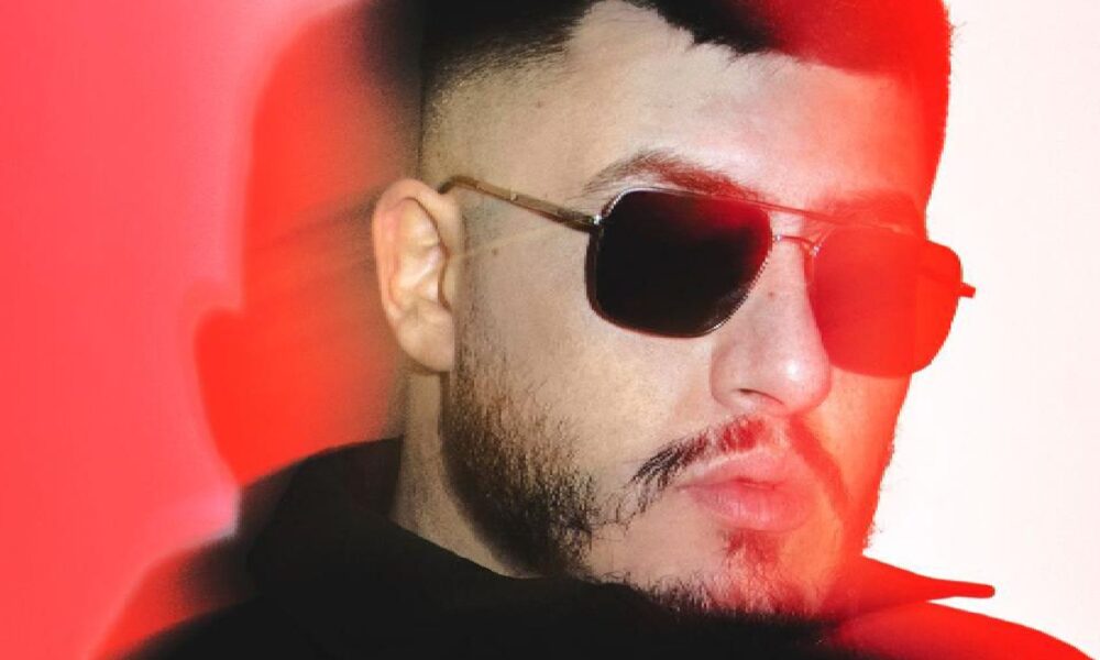 Close-up of a person wearing a black hoodie against a vibrant red and white gradient background with dramatic lighting effects.”