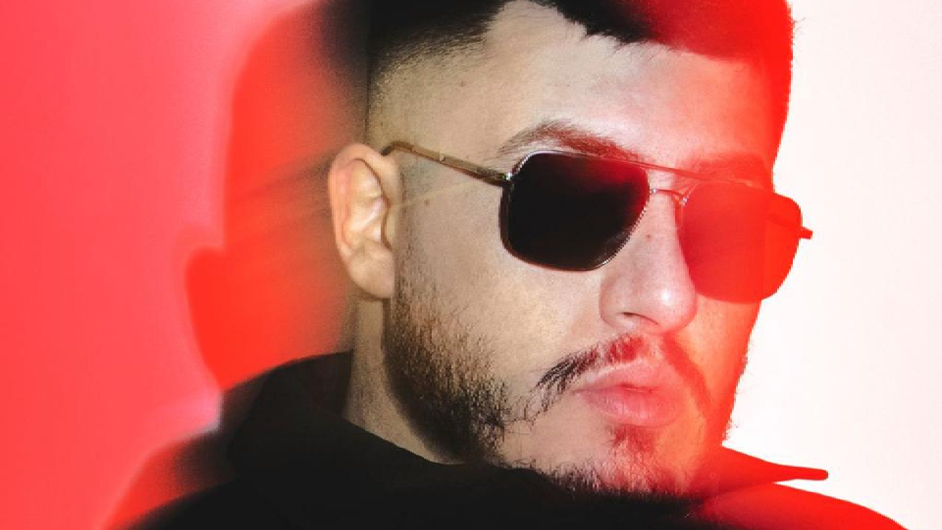 Close-up of a person wearing a black hoodie against a vibrant red and white gradient background with dramatic lighting effects.”