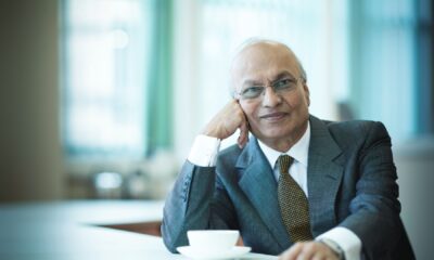 A professional, medium-shot portrait of Mr. Nirmal Sethia, Chairman of Newby Teas. He is an elderly gentleman with distinguished white hair and glasses, dressed in a sharp navy blue blazer, a crisp white shirt, and a patterned silk tie. He is seated in a refined, classic interior—likely a study or private library—with a soft-focus background that emphasizes his thoughtful and authoritative expression as a leader in the luxury tea industry