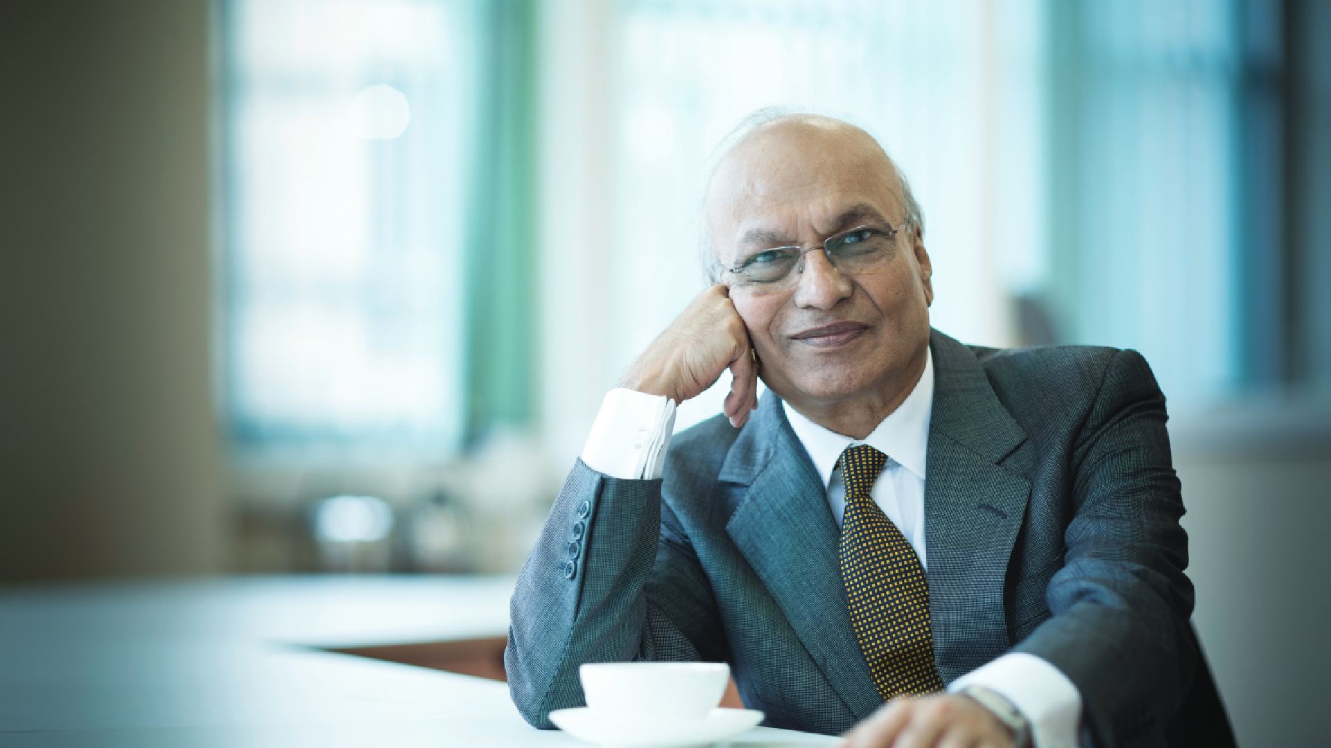 A professional, medium-shot portrait of Mr. Nirmal Sethia, Chairman of Newby Teas. He is an elderly gentleman with distinguished white hair and glasses, dressed in a sharp navy blue blazer, a crisp white shirt, and a patterned silk tie. He is seated in a refined, classic interior—likely a study or private library—with a soft-focus background that emphasizes his thoughtful and authoritative expression as a leader in the luxury tea industry
