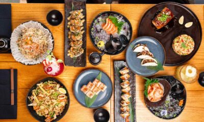 The newly renovated interior of Sushi Art JBR, featuring a design by Quadrant Design that blends Japanese minimalism with natural elements like floating ceiling lights, warm wood textures, and lush greenery. In the foreground, a beautifully plated selection of artisanal sushi, including signature handrolls and Yuzu Miso Marinated Salmon, is presented on a sleek table, capturing the refined and artistic dining atmosphere of the Jumeirah Beach Residence location