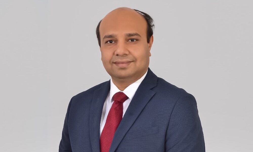 Person wearing a dark blue business suit with a white dress shirt and a red tie, standing against a plain light gray background.