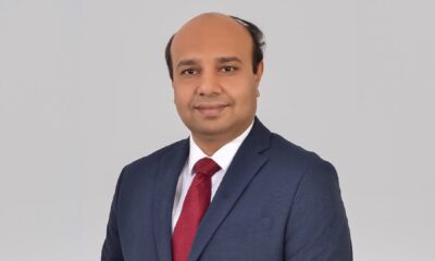 Person wearing a dark blue business suit with a white dress shirt and a red tie, standing against a plain light gray background.