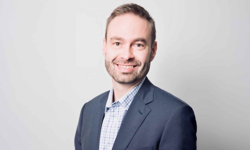 Person wearing a dark business suit and light checkered dress shirt, standing against a plain light gray background.