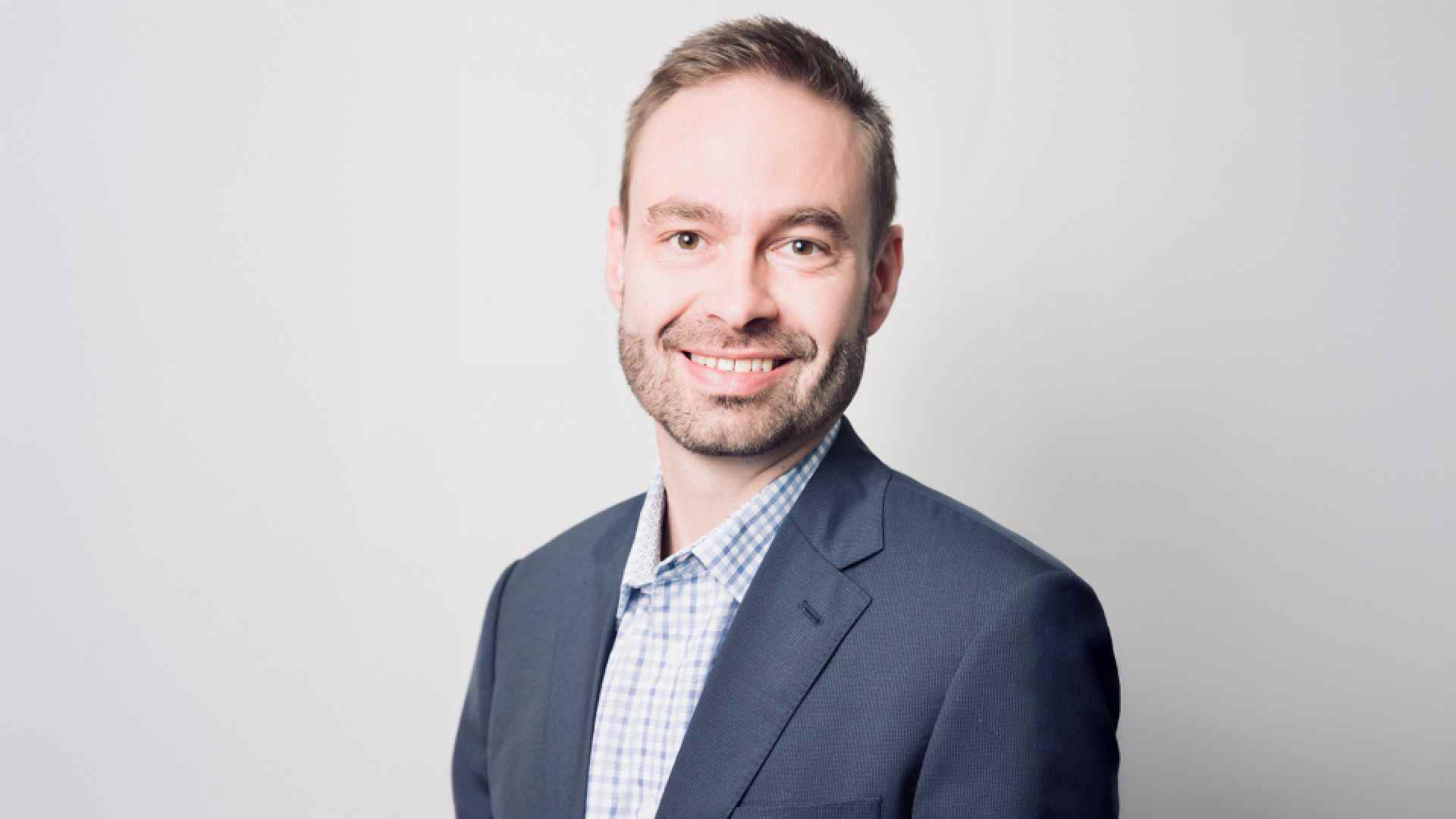 Person wearing a dark business suit and light checkered dress shirt, standing against a plain light gray background.