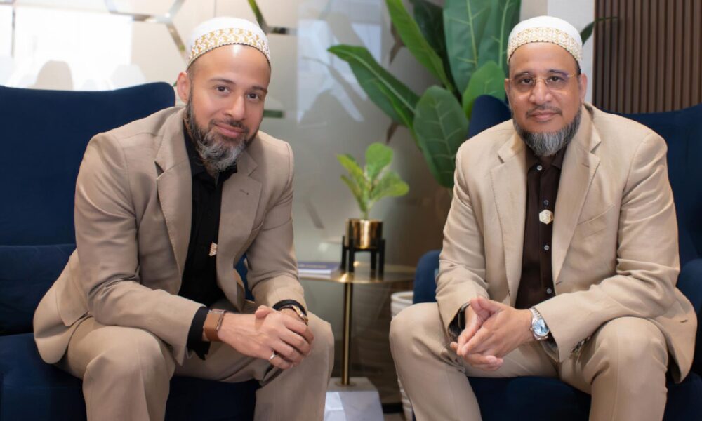 Two individuals seated on dark blue armchairs in a modern indoor setting, wearing beige suits and white caps, with a small gold-accented side table and green potted plant in the background.