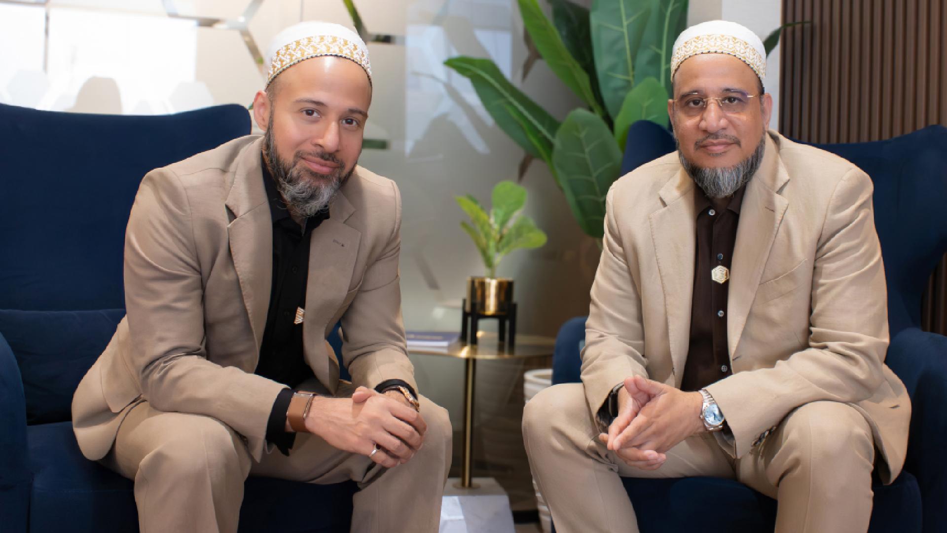 Two individuals seated on dark blue armchairs in a modern indoor setting, wearing beige suits and white caps, with a small gold-accented side table and green potted plant in the background.