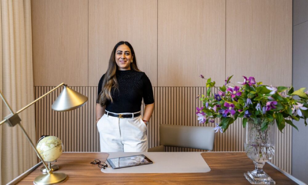 A professional portrait of an architectural or interior designer, likely the founder of KAD Designs, dressed in a business suit and standing in a modern, well-lit interior space.