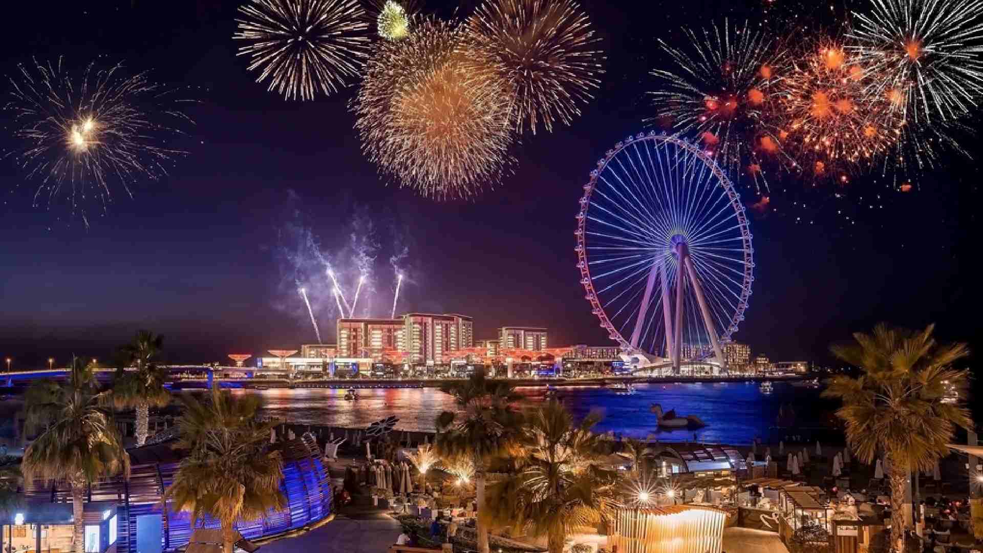Night view of JBR Beach in Dubai with colorful fireworks lighting up the sky above Ain Dubai Ferris wheel, illuminated waterfront buildings, and palm trees in the foreground.