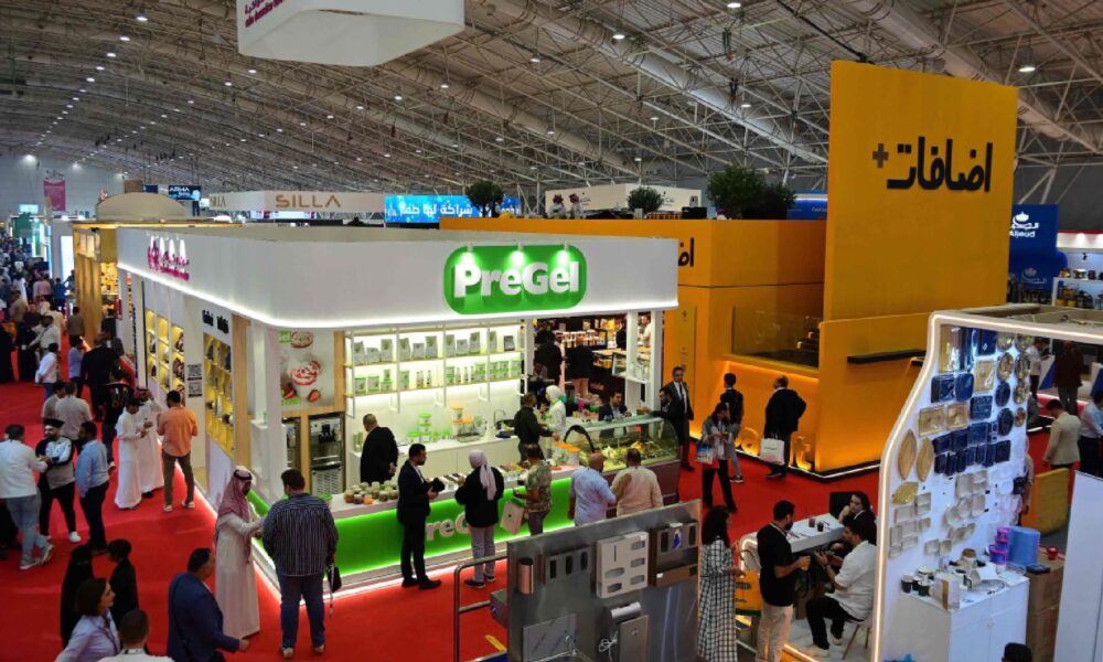 The brightly lit interior of a professional kitchen, featuring chefs gathered around a cooking station during a culinary competition.
