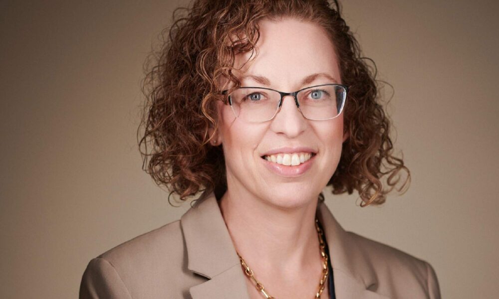 A person wearing a beige blazer and a gold chain necklace against a plain, neutral background.