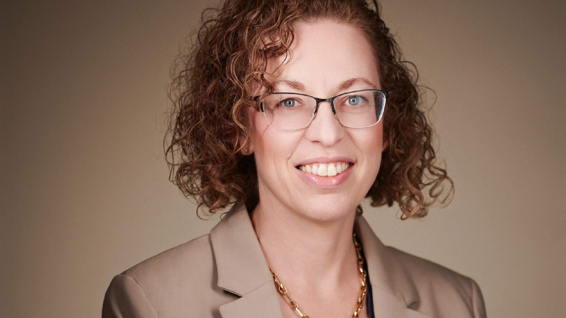 A person wearing a beige blazer and a gold chain necklace against a plain, neutral background.