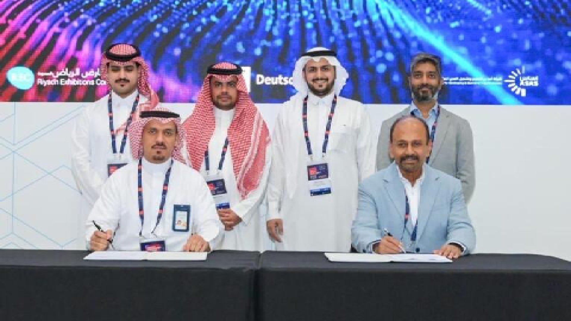 Signing ceremony of a Memorandum of Understanding between Rqeem for Smart Solutions and Torry Harris Business Solutions, with representatives seated at a table signing documents and others standing behind, against a backdrop featuring event branding and digital graphics
