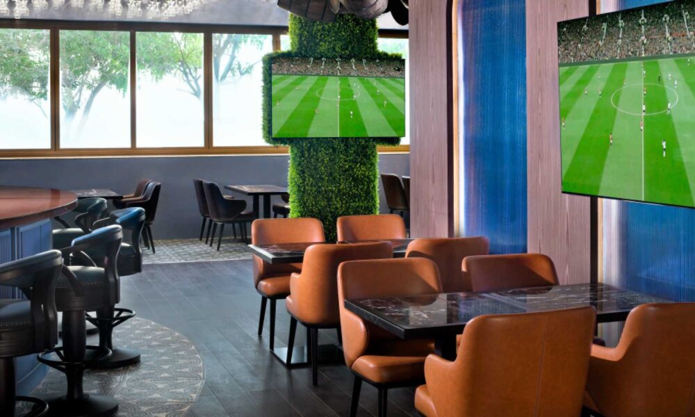 The modern interior of Goose & Gander gastropub in Dubai, featuring tan leather armchairs, marble-topped tables, and multiple flat-screen TVs broadcasting a football match amidst a decor of greenery-covered pillars and decorative wooden barrels