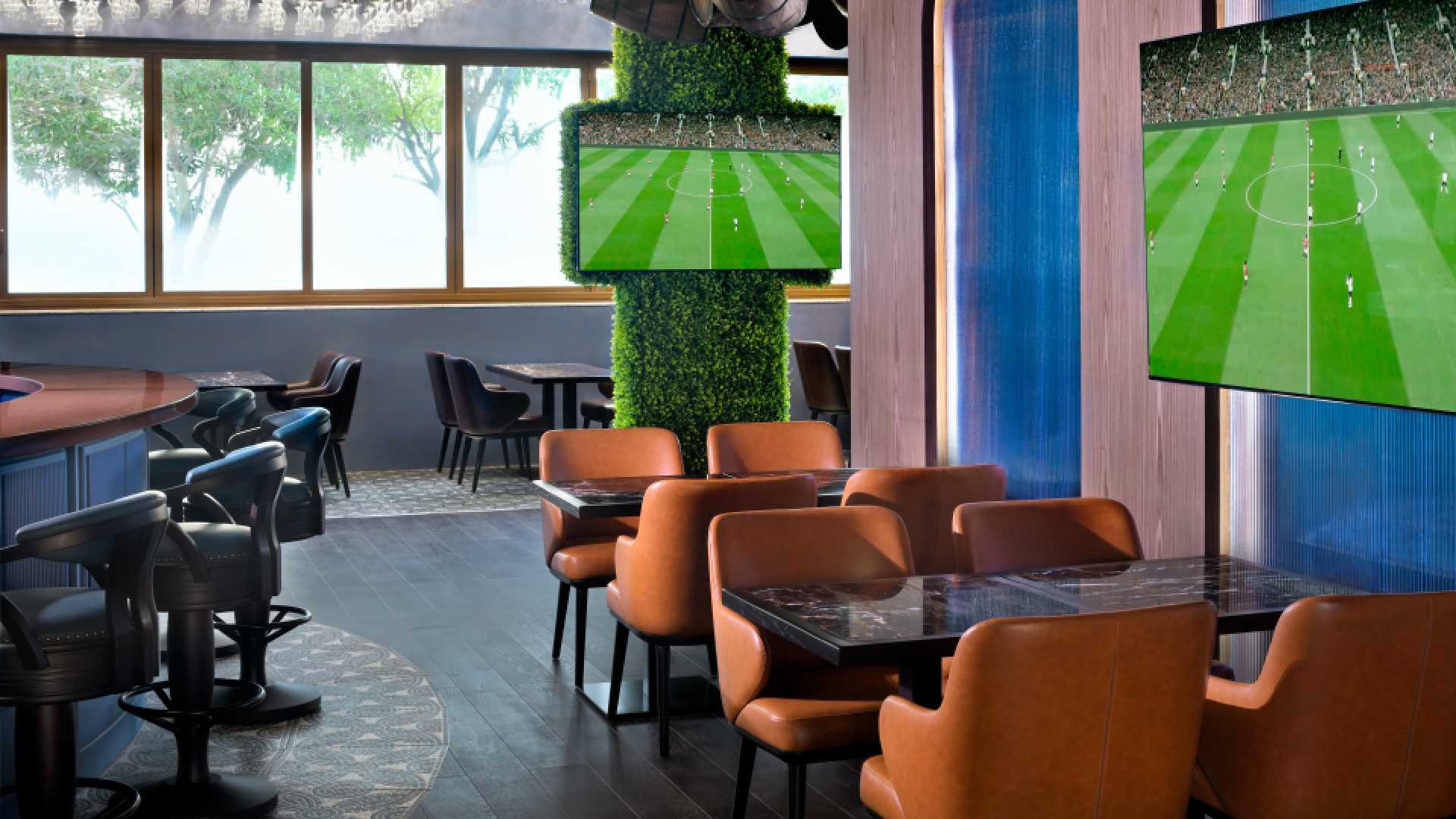 The modern interior of Goose & Gander gastropub in Dubai, featuring tan leather armchairs, marble-topped tables, and multiple flat-screen TVs broadcasting a football match amidst a decor of greenery-covered pillars and decorative wooden barrels