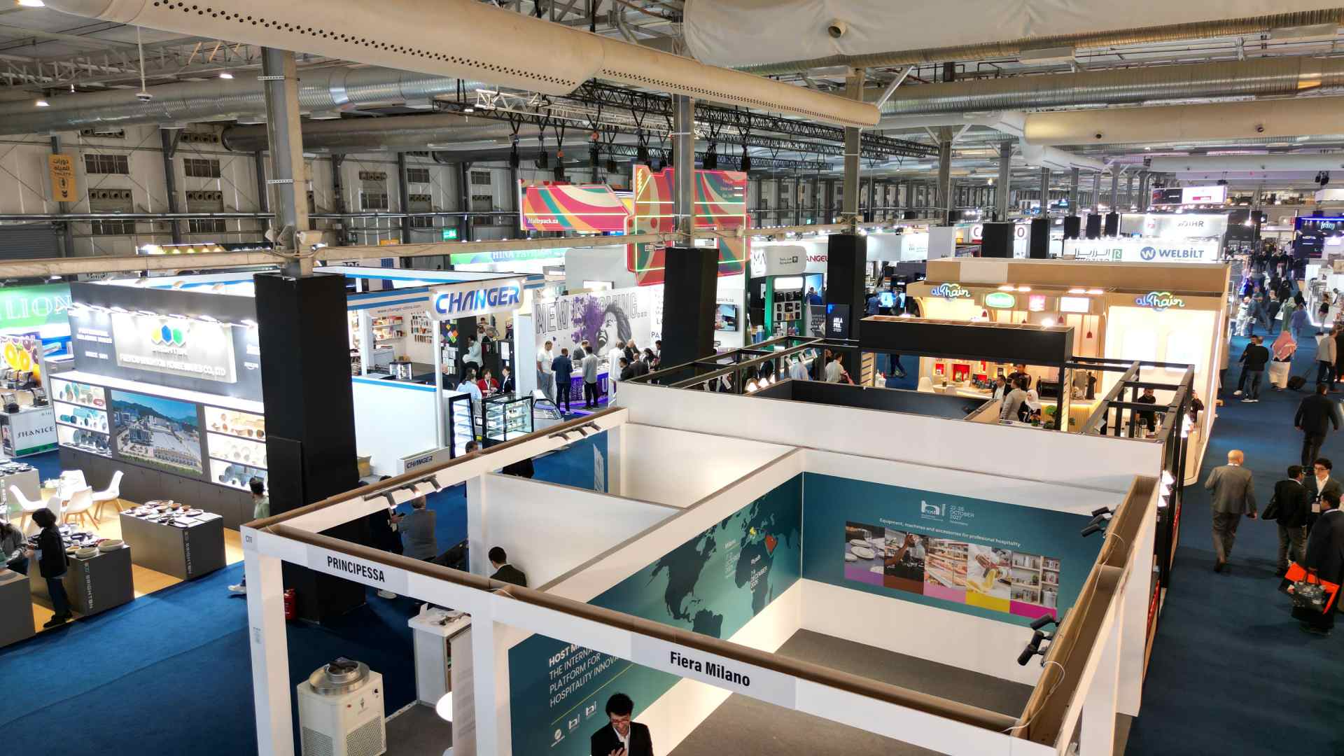 A high-angle, wide-shot of the bustling HORECA Riyadh 2025 exhibition floor at the Riyadh Front Exhibition & Conference Center, showing thousands of professional visitors navigating through 42,000 square meters of sleek corporate booths and hospitality displays under bright industrial lighting