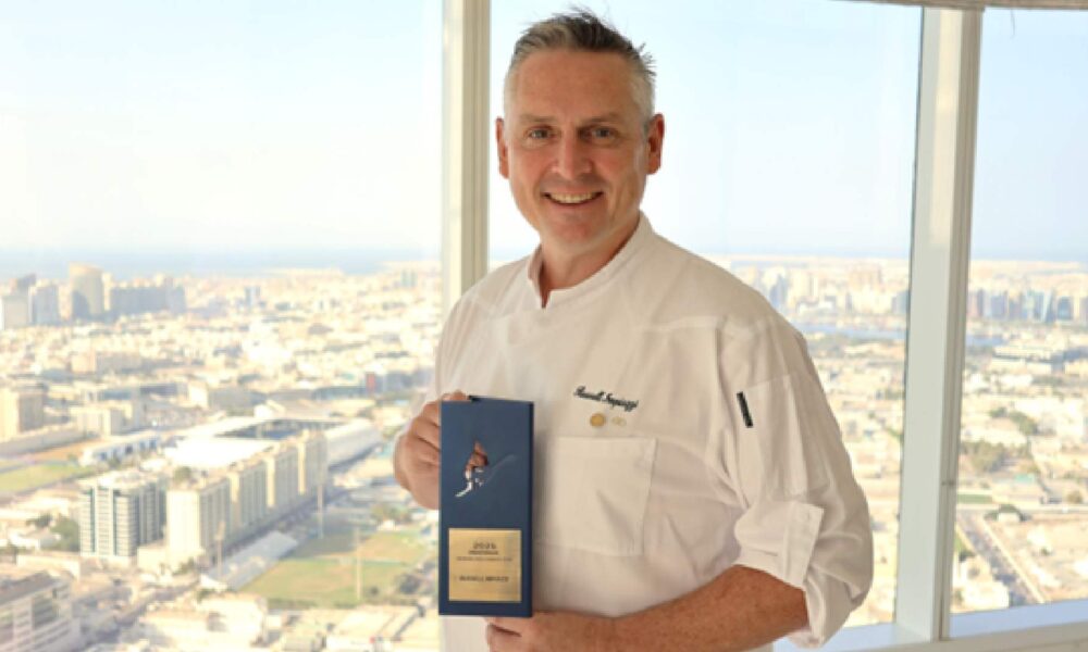 Chef holding an award in front of a large window overlooking a city skyline.
