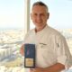 Chef holding an award in front of a large window overlooking a city skyline.