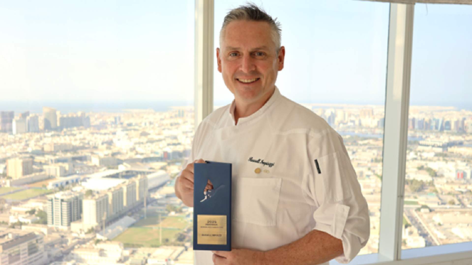 Chef holding an award in front of a large window overlooking a city skyline.