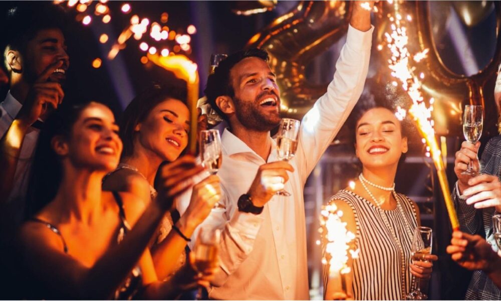 Group of people celebrating and cheering at a party, holding glasses of champagne while sparklers go off in the foreground.