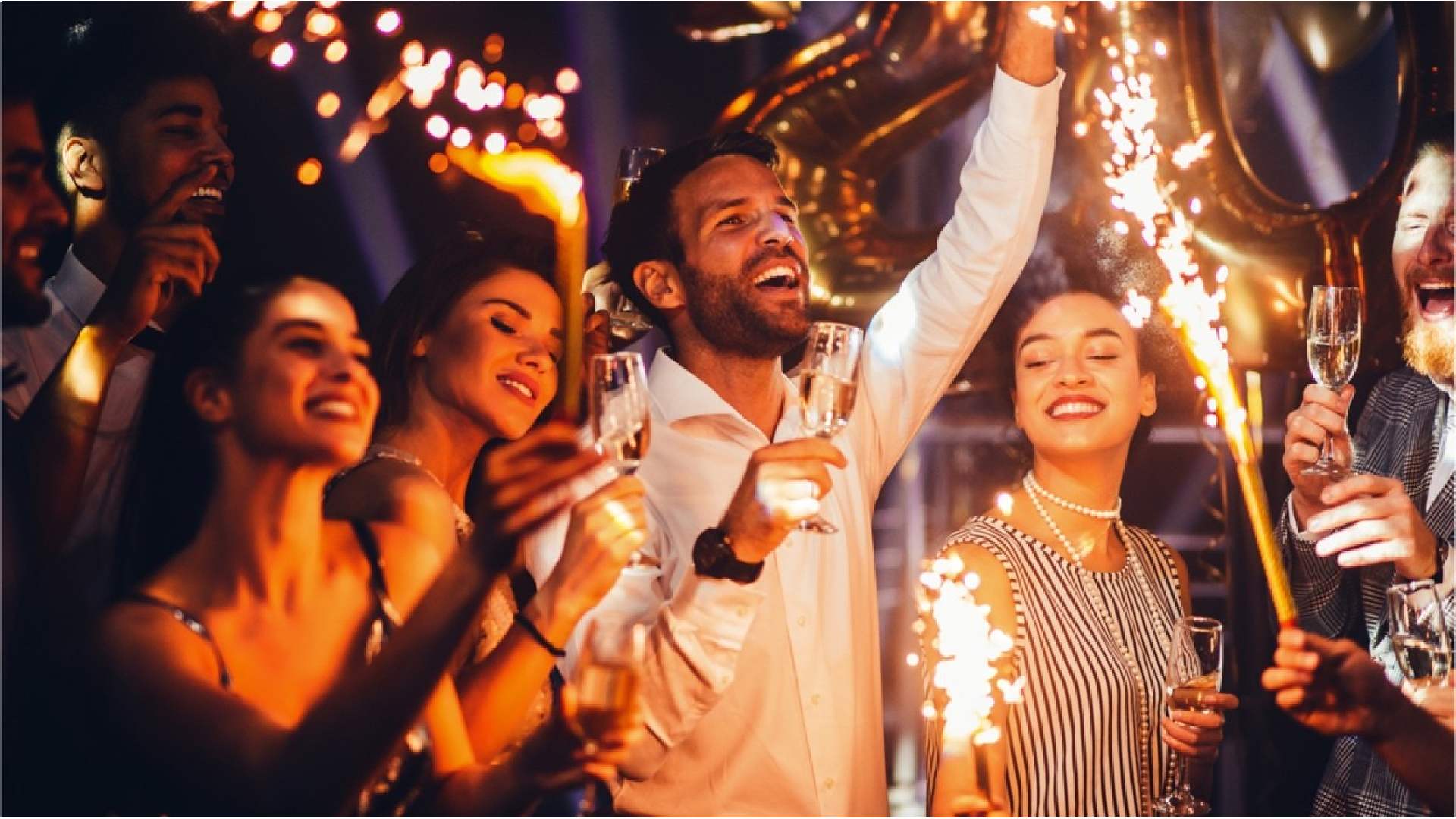 Group of people celebrating and cheering at a party, holding glasses of champagne while sparklers go off in the foreground.