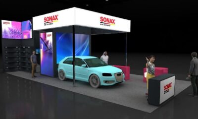 A light blue car displayed under a white canopy at the modern, brightly lit SONAX exhibition booth inside a trade show.