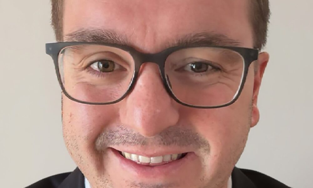 A professional, middle-aged man with dark hair and a beard, wearing a white collared shirt, is seated in a sunlit modern office, smiling directly at the camera.