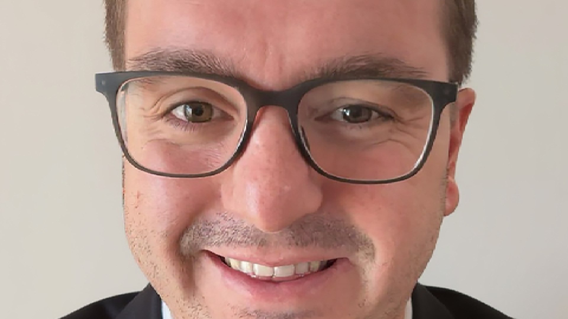 A professional, middle-aged man with dark hair and a beard, wearing a white collared shirt, is seated in a sunlit modern office, smiling directly at the camera.