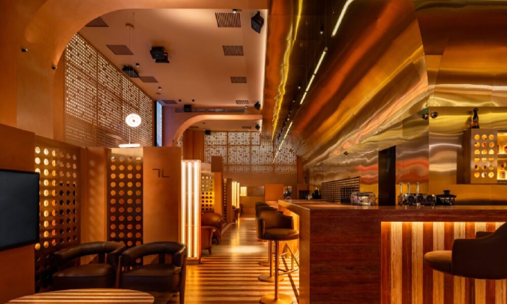 A man wearing glasses and a formal jacket is speaking while seated on a stage, addressing an audience in a large auditorium or conference setting.