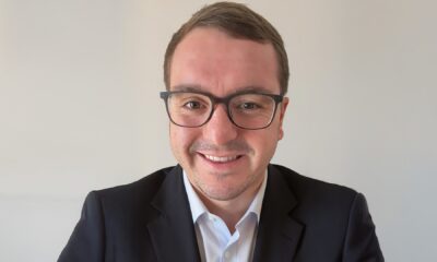 A professional, middle-aged man with dark hair and a beard, wearing a white collared shirt, is seated in a sunlit modern office, smiling directly at the camera.