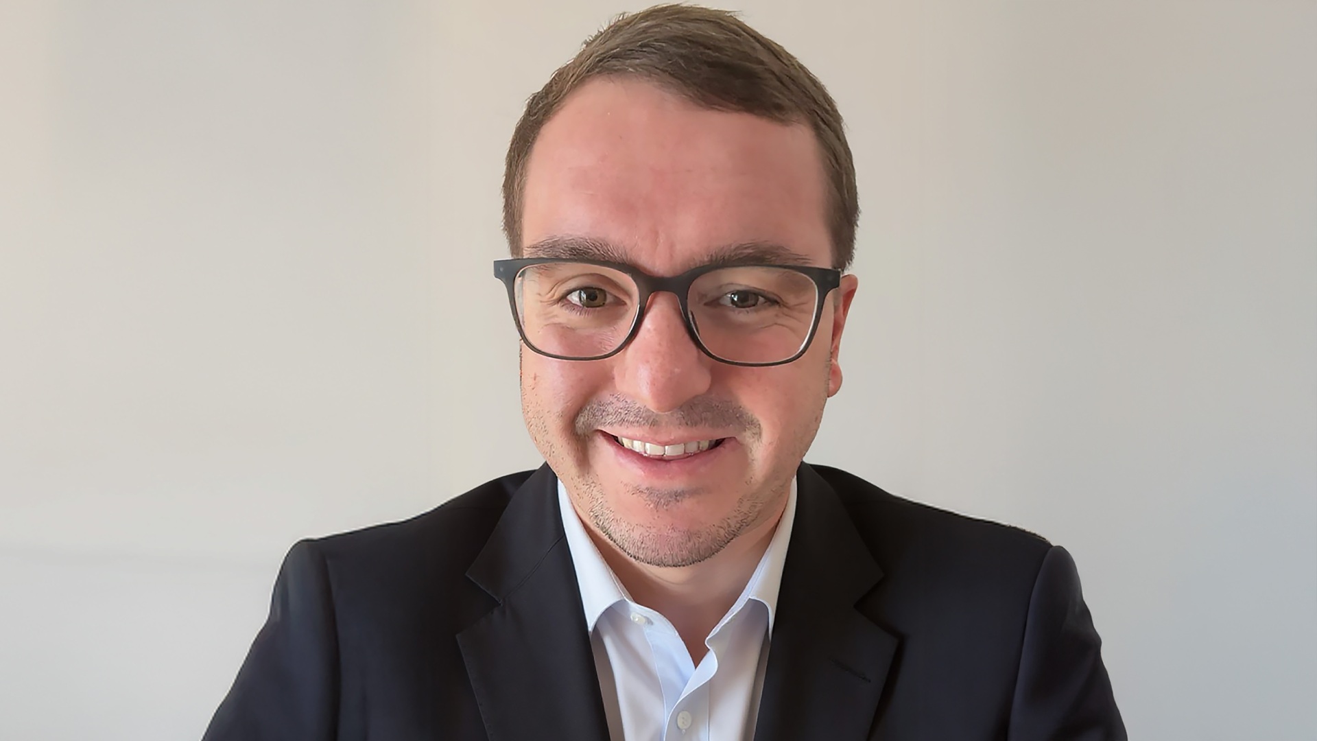 A professional, middle-aged man with dark hair and a beard, wearing a white collared shirt, is seated in a sunlit modern office, smiling directly at the camera.