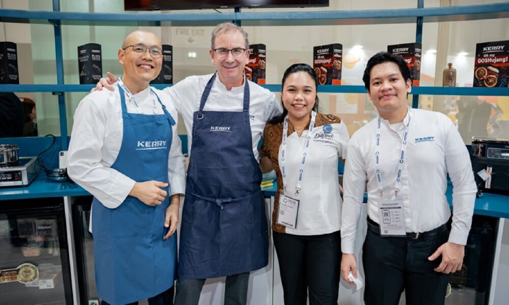 A professional man in a business suit is seated outdoors at a large table under a white tent, looking intently at a presentation screen while a smaller group of people are gathered nearby for a meeting or event.
