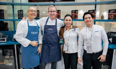 A professional man in a business suit is seated outdoors at a large table under a white tent, looking intently at a presentation screen while a smaller group of people are gathered nearby for a meeting or event.