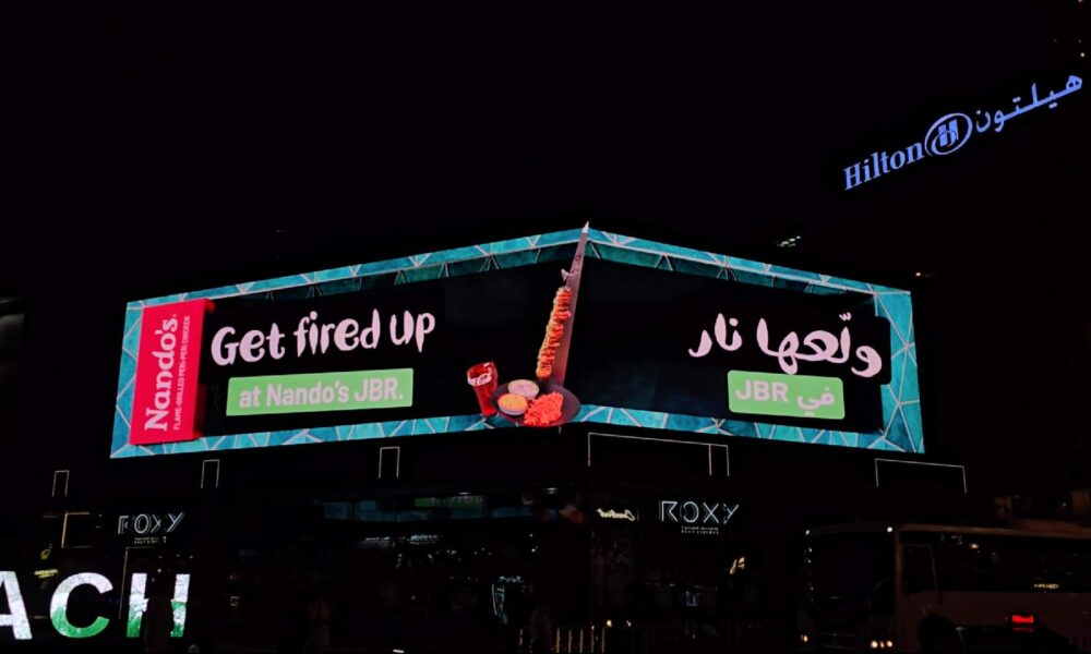 A night photograph of the first-ever global 3D anamorphic billboard for Nando’s, located above Roxy Cinema at The Beach, JBR, in Dubai. The L-shaped display features a hyper-real 3D animation of the new Espetada A Torre Dukkah Lima skewer, a side dish, and a drink appearing to pop out from the screen against a dark background with teal geometric patterns. The billboard prominently displays the slogan 'Get fired up at Nando’s JBR' in both English and Arabic