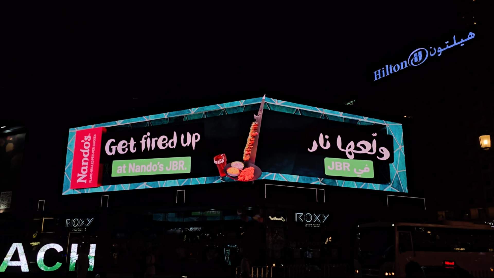 A night photograph of the first-ever global 3D anamorphic billboard for Nando’s, located above Roxy Cinema at The Beach, JBR, in Dubai. The L-shaped display features a hyper-real 3D animation of the new Espetada A Torre Dukkah Lima skewer, a side dish, and a drink appearing to pop out from the screen against a dark background with teal geometric patterns. The billboard prominently displays the slogan 'Get fired up at Nando’s JBR' in both English and Arabic