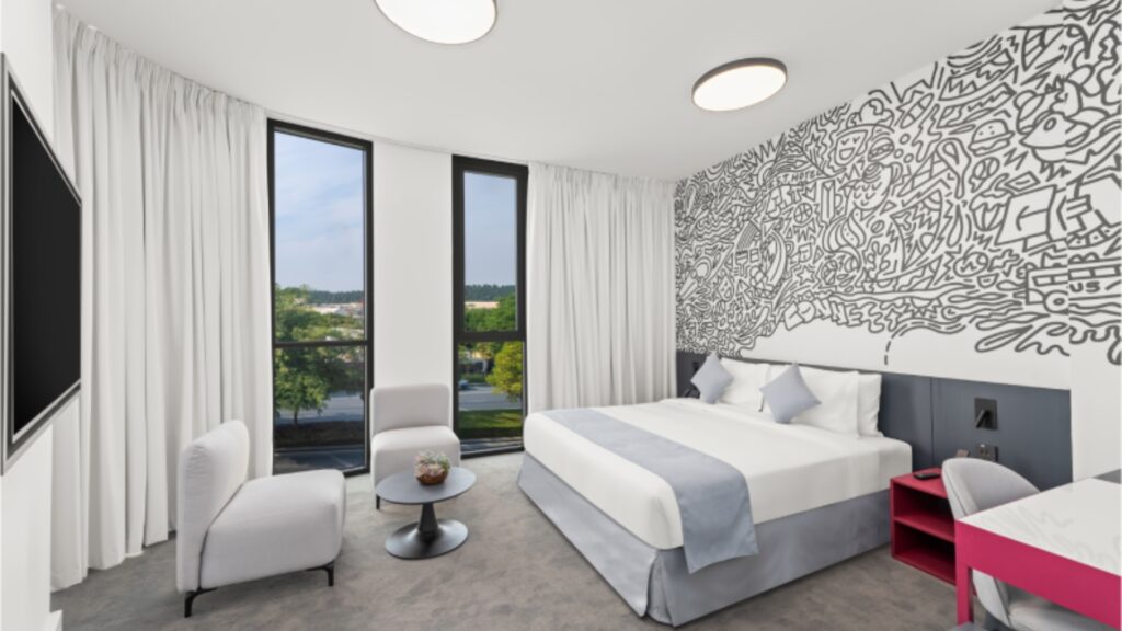 A contemporary hotel room featuring a queen or king-sized bed with white linens and a light blue accent runner. The wall behind the bed has a large, black and white graffiti-style mural. To the left, two modern white lounge chairs sit near floor-to-ceiling windows draped with white curtains, offering a view of green trees outside.
