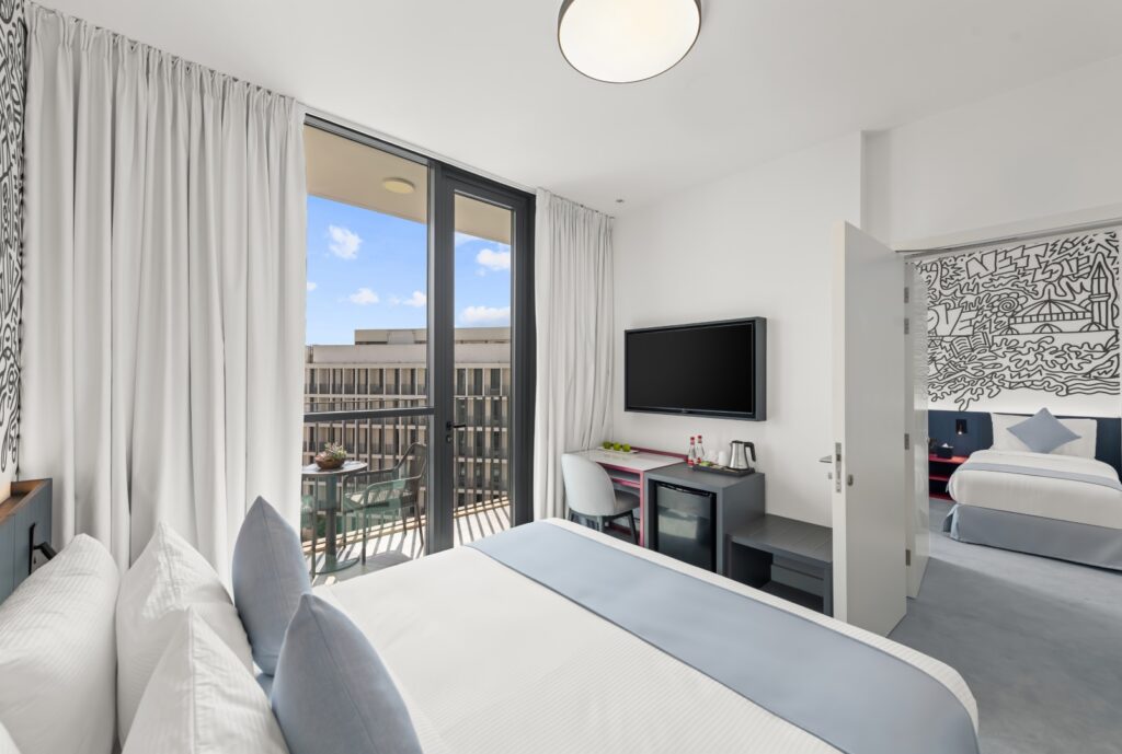 A bright hotel room featuring a large bed with white linens and light blue pillows. A TV is mounted on the wall above a small desk/minibar area. A glass sliding door leads out to a balcony with outdoor seating (visible from the first image). Through an open doorway on the right, a second smaller room with another bed is visible, with a black and white graffiti-style mural on the far wall.