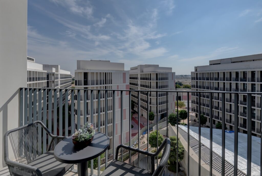 A view from a balcony showing two dark wicker chairs and a small table with a floral centerpiece. The balcony overlooks a complex of modern, light-colored apartment buildings with black railings under a bright, partly cloudy sky.