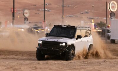 A white Jetour T2 off-road vehicle kicks up a large cloud of sand as it drives across the desert during the day. The vehicle is modified with a black hood decal and a roof rack. The background shows the hazy outlines of desert structures and festival signage under a sunset sky.